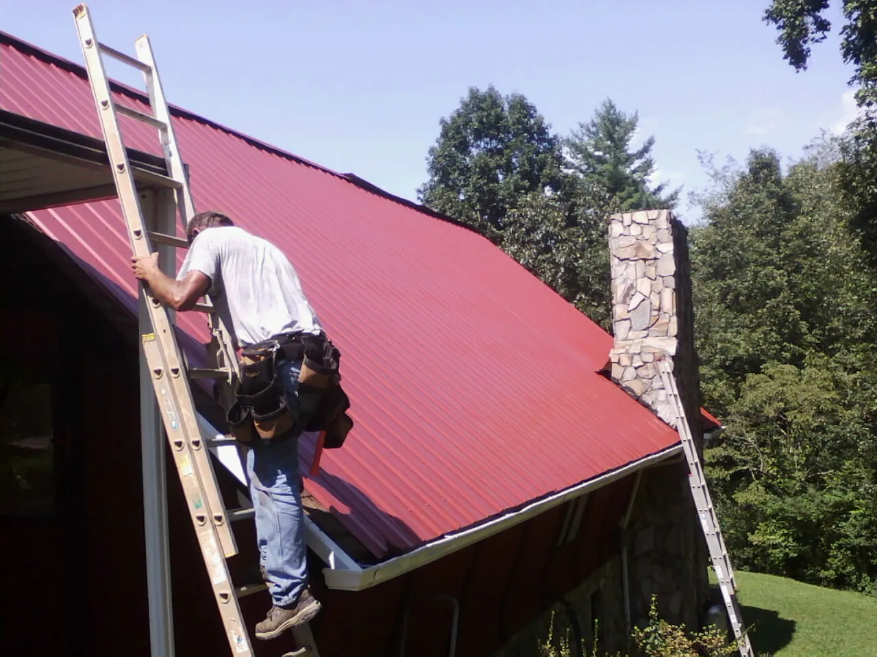 Roofer on a ladder beside a red metal roof and stone chimney — Soffit & Fascia Repair by Roofing Brothers in Blue River
